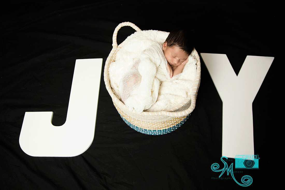 a baby boy sleeps peacefully in a round basket which forms the letter "O" in the word "Joy"