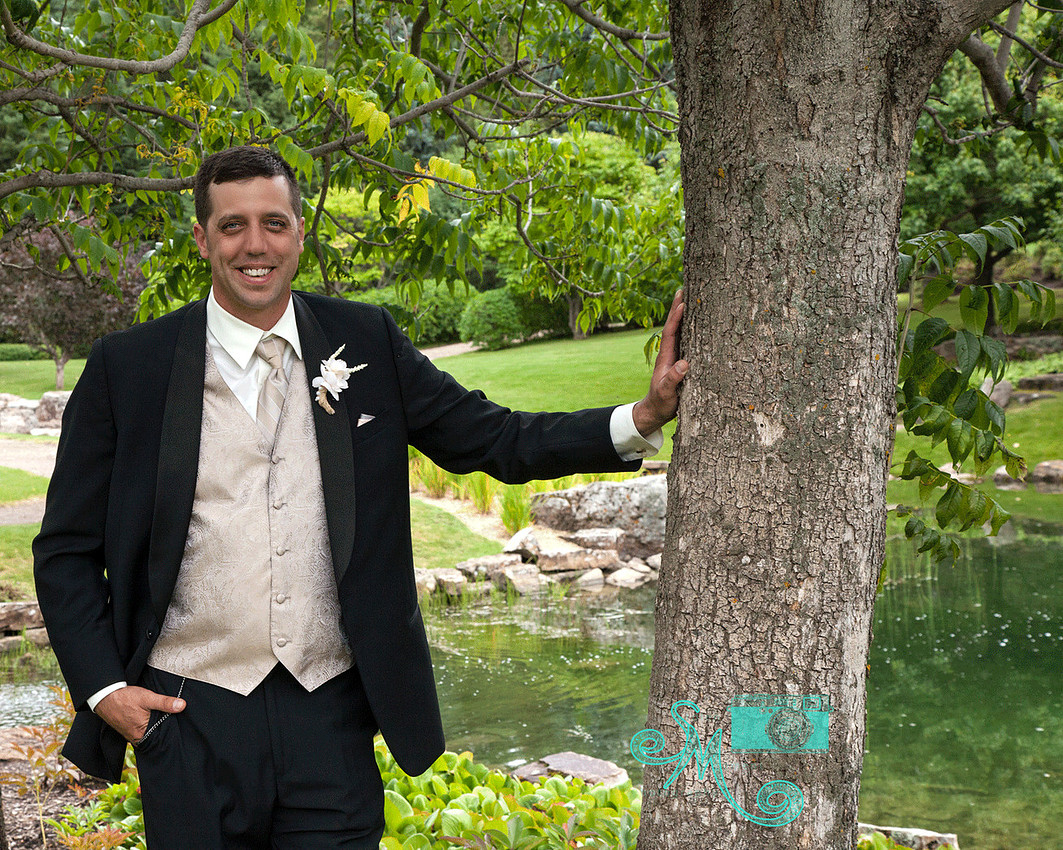 Groom standing with hand on tree with pond in background