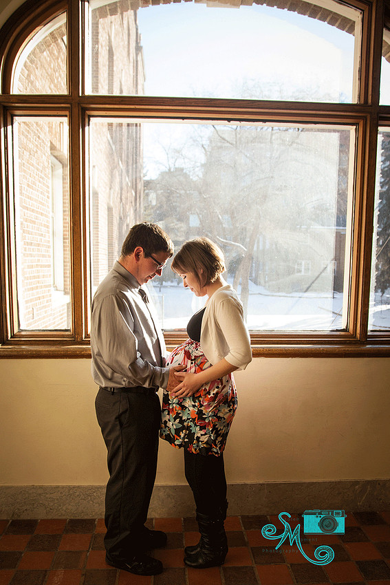 a mom and dad to be hold the belly while standing in front of a large window with the sun pouring in