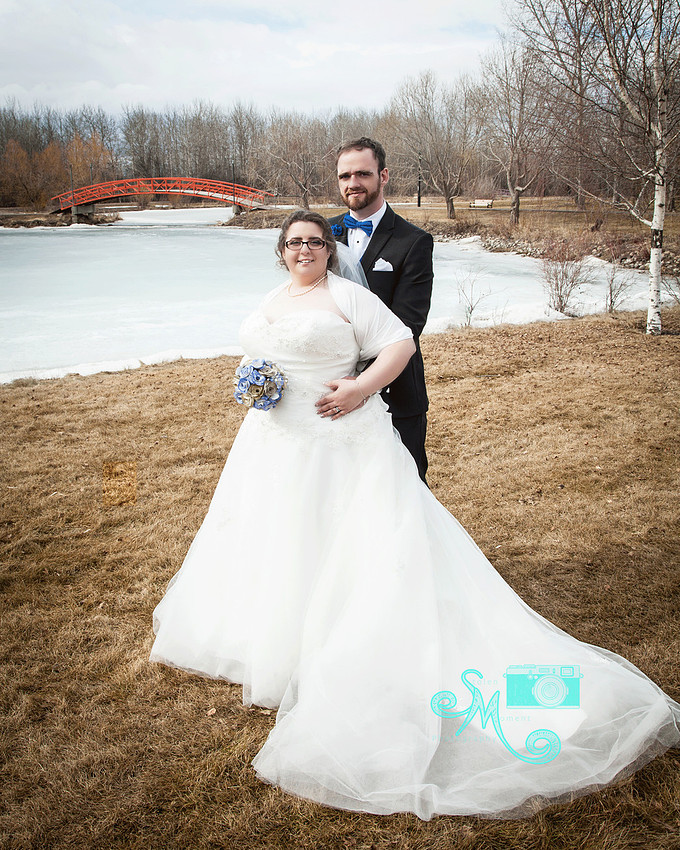 bride and groom pose for formal photo at dog rump creek park with bridge and lake in background