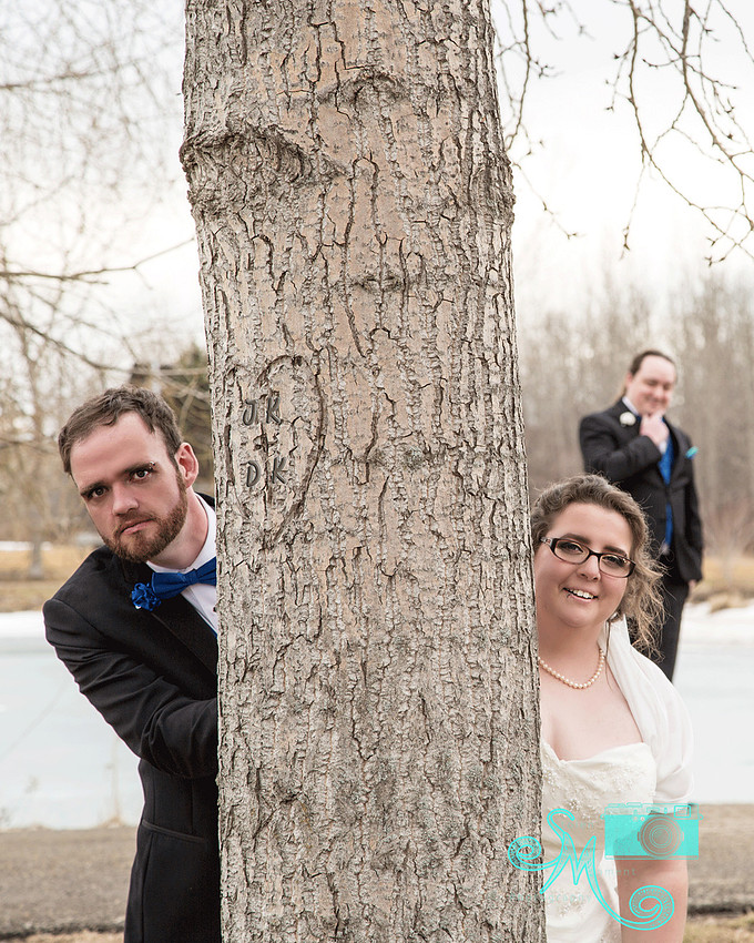 bride and groom peeking around a tree while a groomsmen looks on