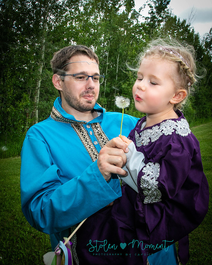 the bride's niece is held in her father's arms both dressed in fourteenth century costume and she blows dandelion fuzz