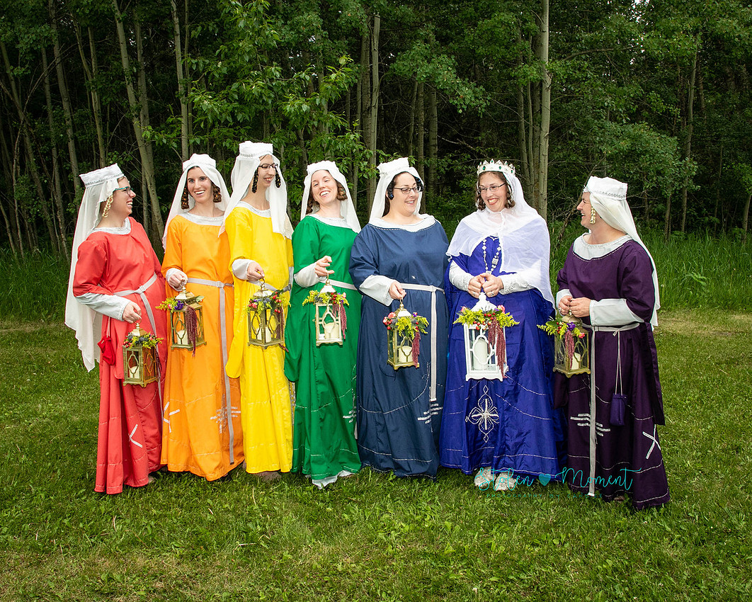 the bride and her maids stand in a line in traditional medieval costume holding lanterns and sharing a laugh between themselves
