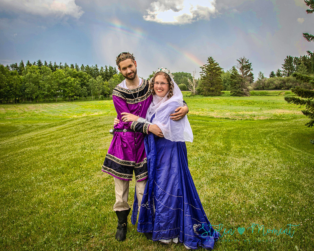 a bride and groom in each other's arms smiling at the camera both dressed in medieval costumes with a rainbow in the sky behind them