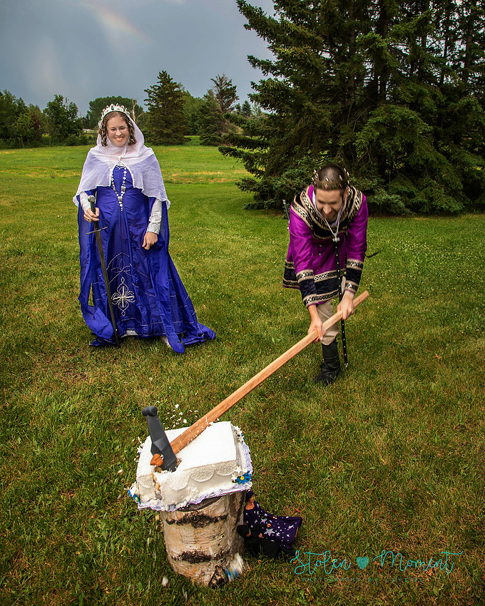 the groom destroys the wedding cake with a medieval style weapon while the bride looks on and smiles