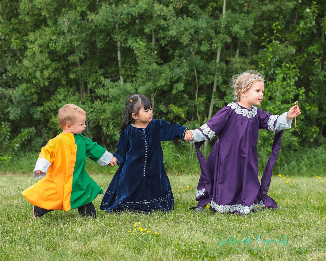 three children from a medieval wedding, all dressed in traditional costume hold hands and walk across the grass, with a girl in the lead carrying a dandelion
