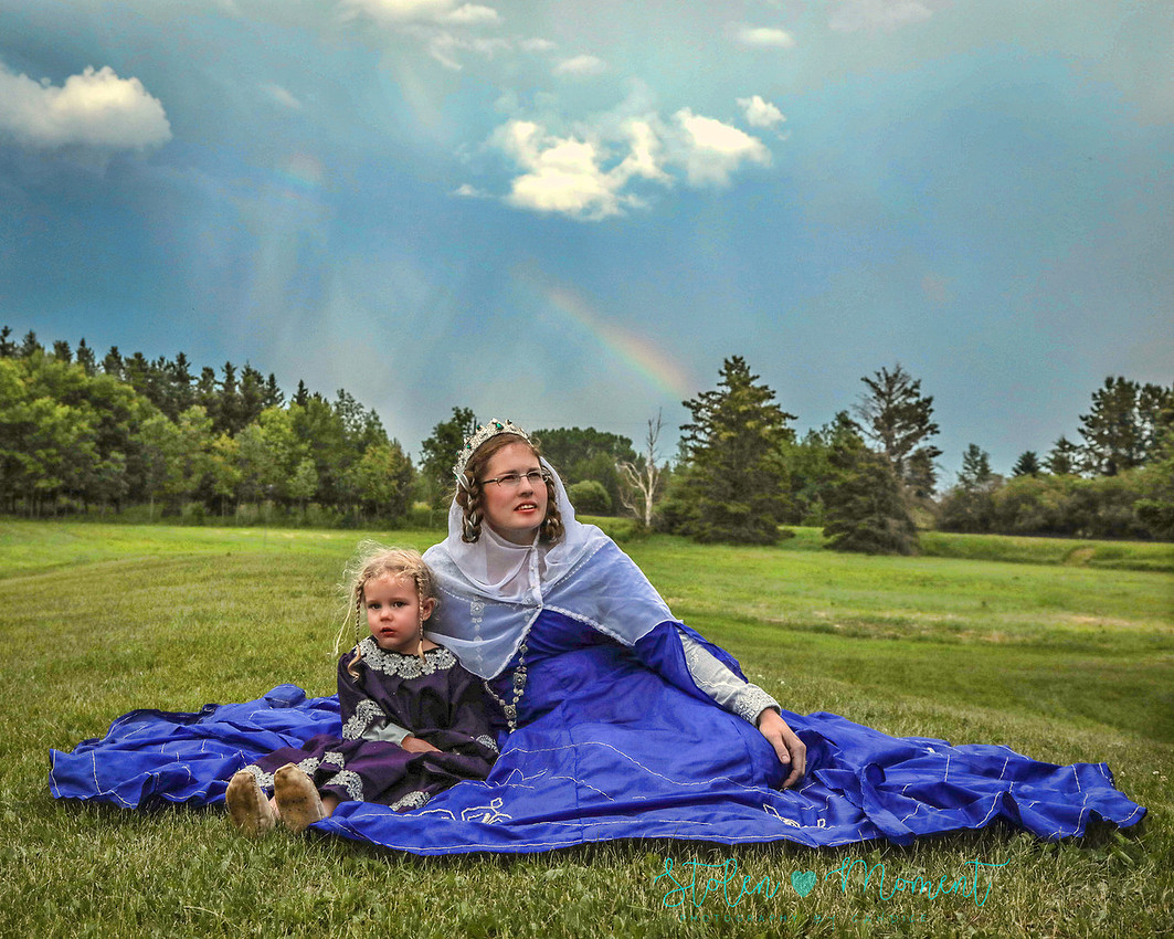 a bride sits on the grass with one of her flower girls beside her both dressed in medieval costumes with a rainbow in the sky behind them