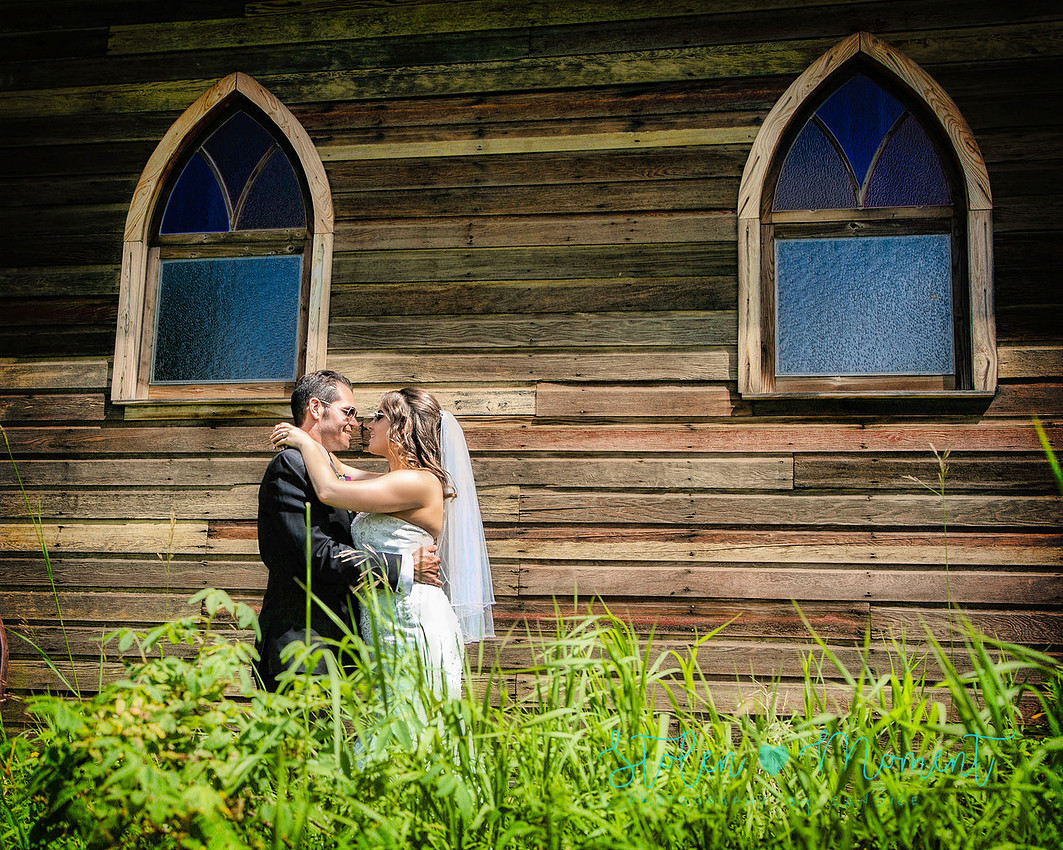 a bride and groom in each others arms looking into each others eyes in front of an old wooden church at Hastings lake Gardens
