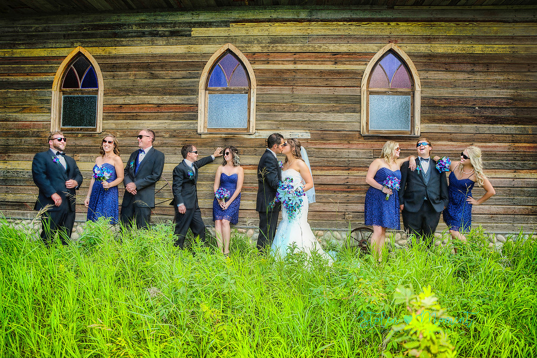 a wedding party of ten stand in front of an old wooden church at Hastings Lake Gardens.  The party share a laugh while the bride and groom kiss