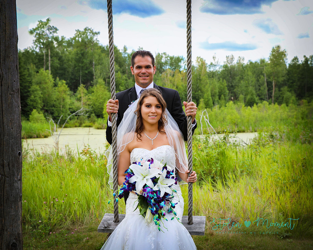 a groom stands behind a swing as his bride sits on the swing at Hastings Lake Gardens