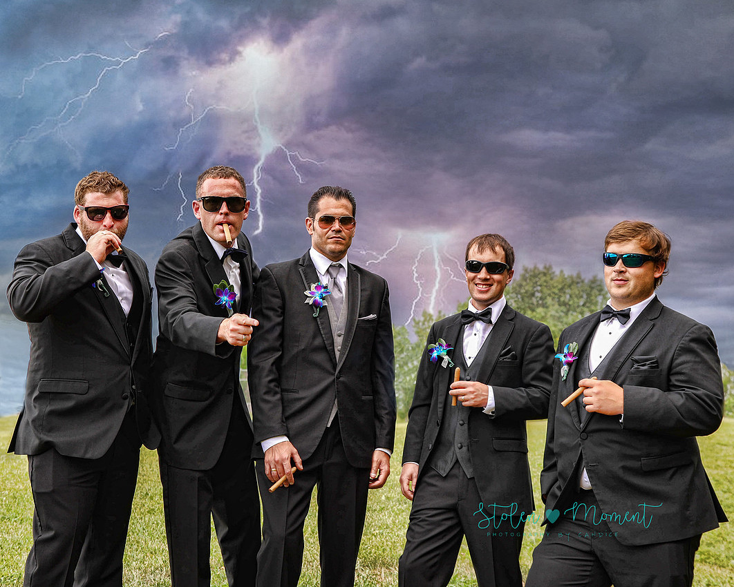 the groom and his four groomsmen all wearing sunglasses and holding cigars pose seriously while the sky is menacing looking overhead