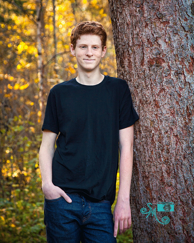 a teenage boy posing against a tree with fall leaves int he background