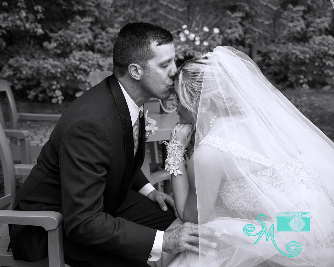 bride and groom sitting across from each other and groom leans in to kiss bride on forehead
