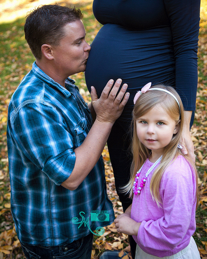  man kisses his pregnant wife's belly while their daughter stands beside them