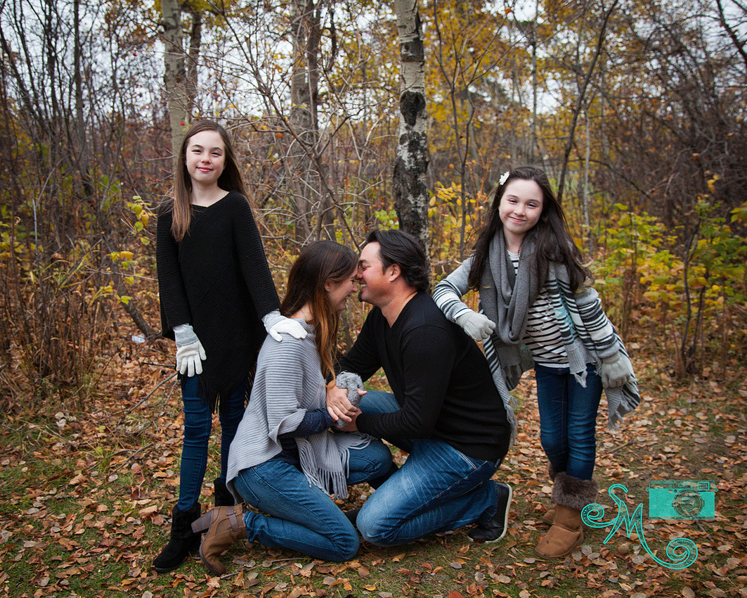 a mom and dad laughing while looking at each other while two girls pose standing beside them in the fall leaves