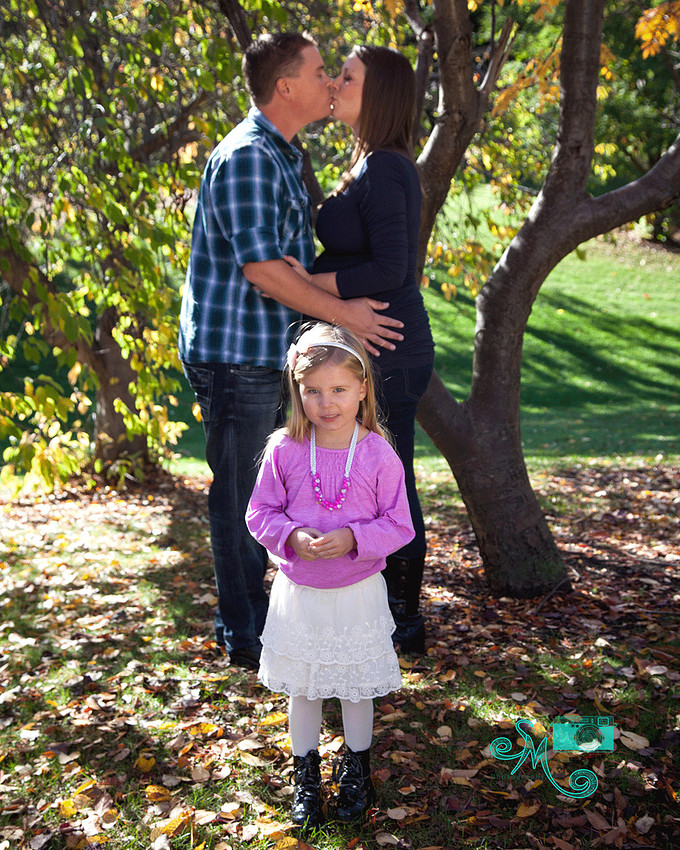 Mom and Dad kiss in the background while a big sister-to-be smiles while standing in front of them 